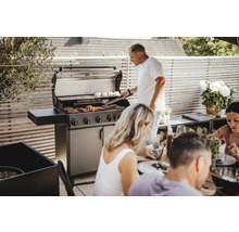 Szene im Freien mit einem Mann, der an einem Gasgrill Essen zubereitet, während eine Gruppe von Menschen an einem Tisch sitzt