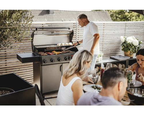 Szene im Freien mit einem Mann, der an einem Gasgrill Essen zubereitet, während eine Gruppe von Menschen an einem Tisch sitzt
