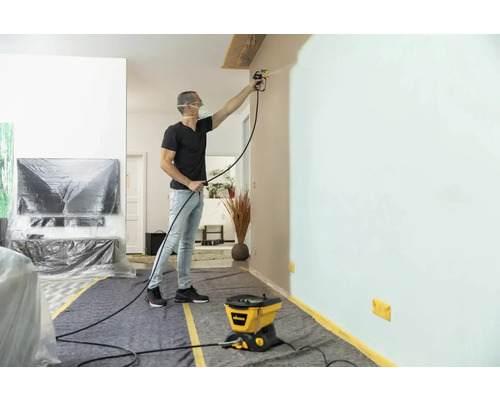 Un homme peint un mur avec un système de pulvérisation de peinture dans un salon protégé par des matériaux de couverture.