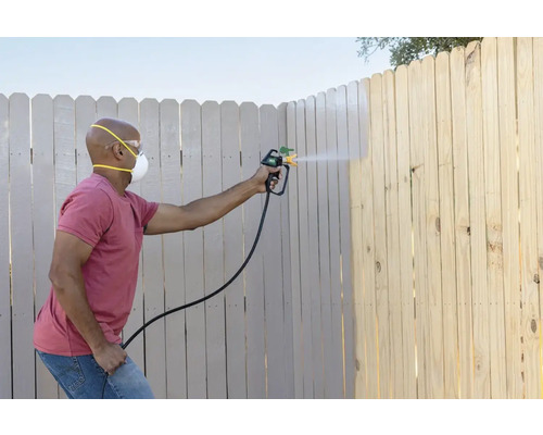 Une personne peint une clôture en bois avec un pistolet à peinture, portant un masque de protection et des lunettes de sécurité.