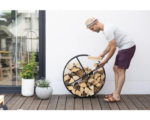 Un homme remplit de bois un range-bûches rond en métal sur une terrasse