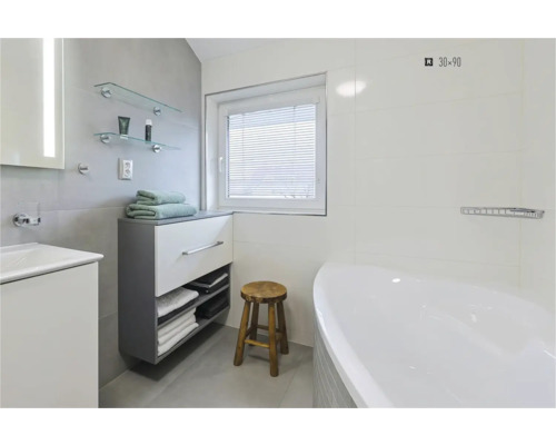 Helles Badezimmer mit Badewanne, Waschtisch, Unterschrank, Handtüchern, Holzschemel und Fenster mit Jalousie