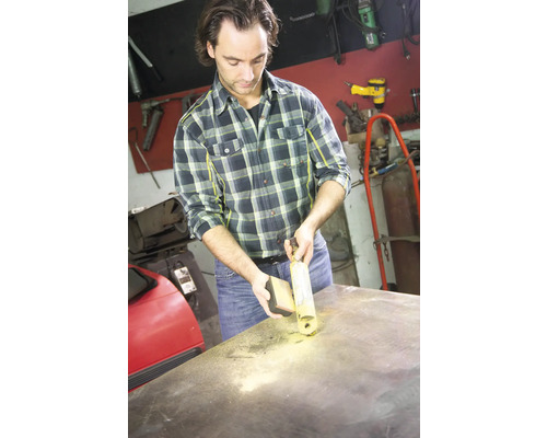 Mann reinigt eine Oberfläche mit einem Reinigungsspray und einem Schwamm in einer Werkstatt.