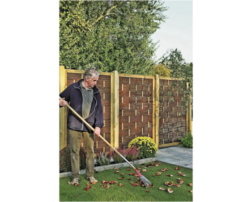 Un homme ramasse les feuilles devant une clôture en bois avec motif tressé et poteaux en bois dans un jardin.
