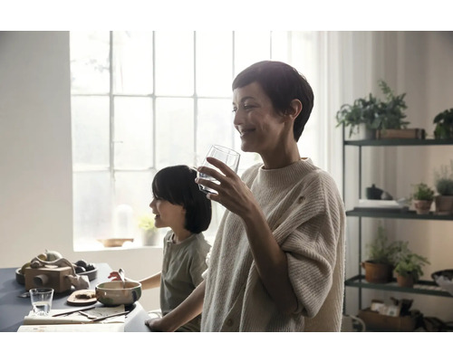Helle Küche mit Mutter und Kind am Tisch, die ein Glas Wasser trinkt. Im Hintergrund befindet sich ein Regal mit Zimmerpflanzen.