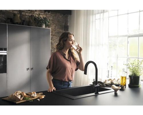 Frau trinkt Wasser in einer hellen Küche mit schwarzem Spülbecken und Armatur
