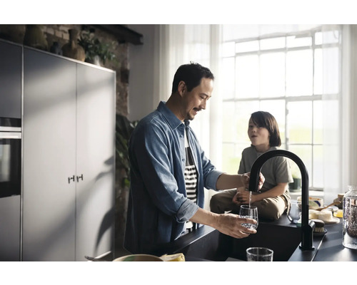 Ein Mann füllt in einer hellen Küche ein Glas mit einem schwarzen Wasserhahn.