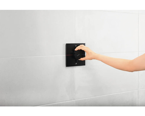 Une main actionne un thermostat de douche noir sur un mur blanc.