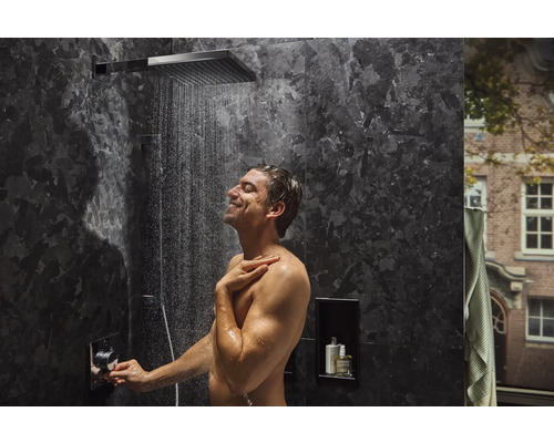 Homme sous un robinet de douche avec pomme de douche plate dans une salle de bain avec des murs en marbre foncé.