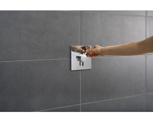 Main actionnant la robinetterie de douche avec façade carrée sur un mur gris