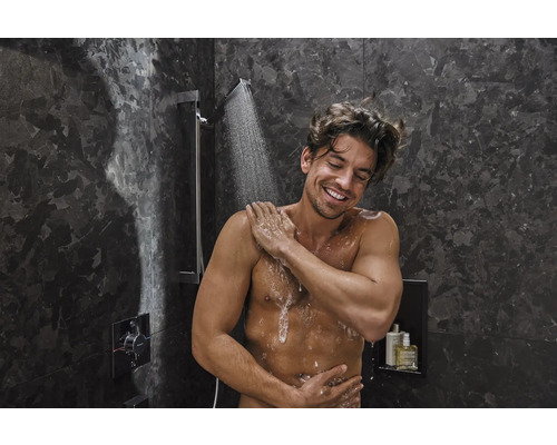 Un homme prend une douche sous un pommeau de douche dans une salle de bains aux murs en marbre foncé.