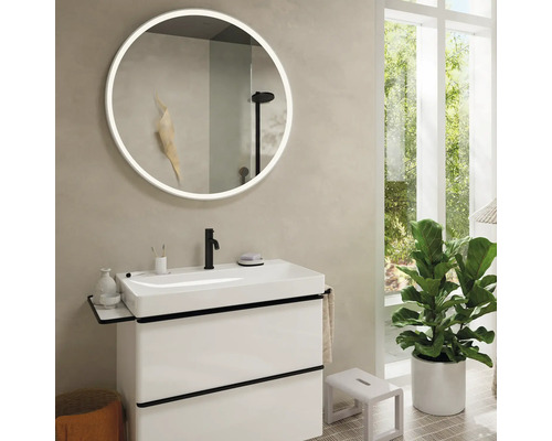 Helles Badezimmer mit Waschtisch, rundem Spiegel mit Beleuchtung und Zimmerpflanze.