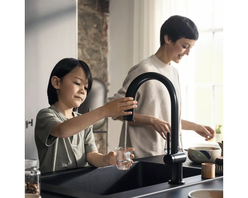 In einer Küche füllt ein Kind Wasser aus einem schwarzen Wasserhahn in ein Glas, während eine Frau im Hintergrund steht.