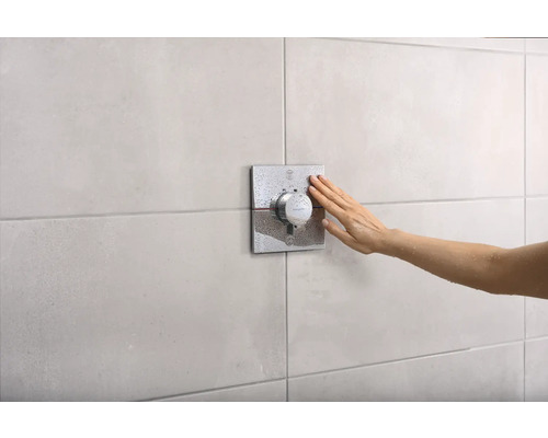 Une main actionne un thermostat de douche sur un mur carrelé