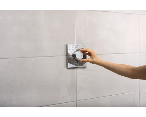 Une main actionne un mitigeur de douche avec une rosace carrée dans une salle de bain.