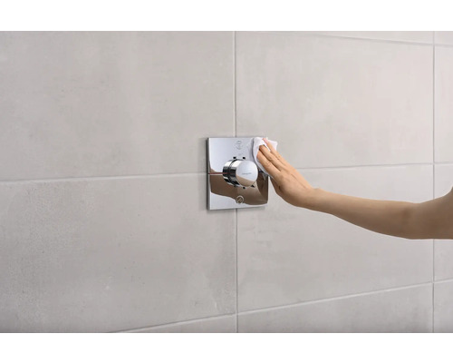 Une main nettoie un thermostat de douche chromé sur le mur.