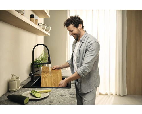 Un homme lave une planche à découper en bois dans un évier de cuisine noir sous un robinet noir.
