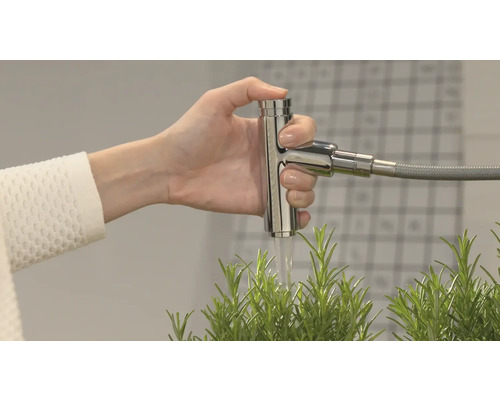 Eine Hand betätigt einen Wasserhahn aus Metall, aus dem Wasser über eine Pflanze fließt.