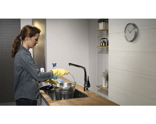Femme portant des gants de ménage remplissant un seau à l''évier dans une cuisine lumineuse.