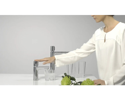 Frau bedient einen Wasserhahn aus Metall in einem hellen Badezimmer.