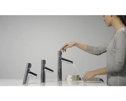 Drei glänzende Waschtischarmaturen unterschiedlicher Höhe, wobei eine Armatur von einer Person bedient wird und Wasser fließt