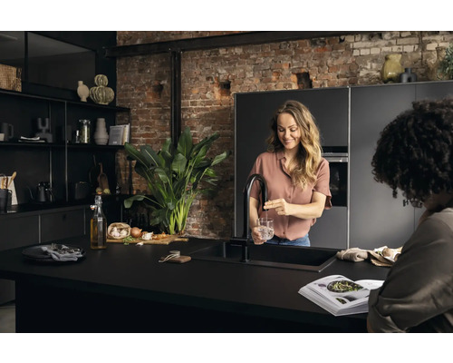 Moderne Küche mit dunklen Möbeln, einer Frau, die Wasser in ein Glas füllt, und einem offenen Kochbuch auf der Arbeitsplatte.
