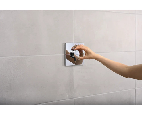 Une main actionne un thermostat carré sur un mur carrelé