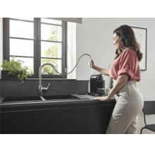 Frau befüllt Wassertank einer schwarzen Kaffeemaschine neben einer Spüle in einer modernen Küche.