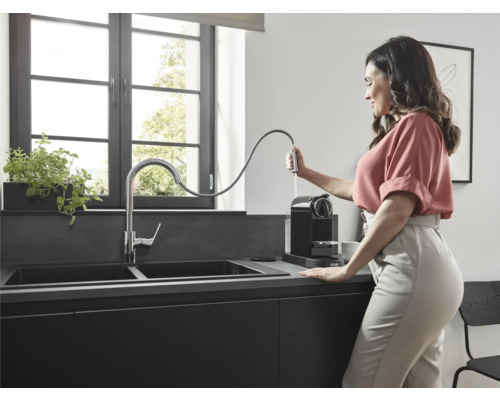 Frau befüllt Wassertank einer schwarzen Kaffeemaschine neben einer Spüle in einer modernen Küche.