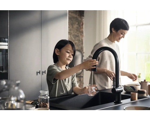 In einer hellen Küche füllt ein Kind ein Glas Wasser an einem schwarzen Wasserhahn, während eine Frau im Hintergrund arbeitet.