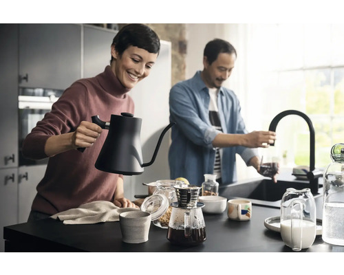 Szene in einer Küche mit zwei Personen, die Kaffee zubereiten, mit einem Wasserkocher, einer Kaffeemaschine und einer Küchenarmatur.