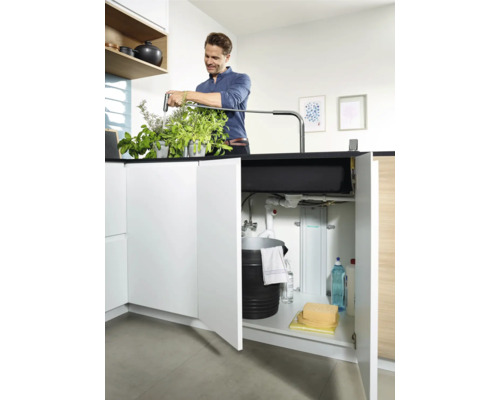 Un homme arrose des herbes aromatiques près de l''évier avec un meuble bas ouvert contenant un seau et des produits de nettoyage.