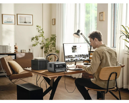 Mann arbeitet an einem Computer mit einem tragbaren Kraftwerk, einem Router und einem Netzwerkspeichergerät auf einem Tisch in einem hellen Raum.