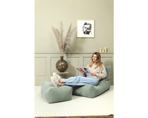 Une femme lit sur une chaise en velours côtelé verte avec un repose-pieds dans une pièce lumineuse.