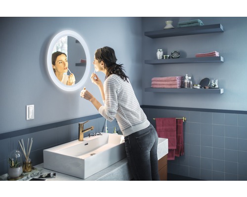 Salle de bains avec miroir lumineux, lavabo et étagères murales. Une femme met du rouge à lèvres.