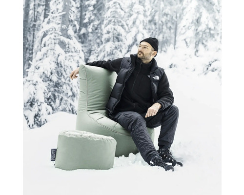 Mann sëtzt an engem grénge Sëtzsack mat Fousshocker am Schnéi virun engem verschneite Bësch