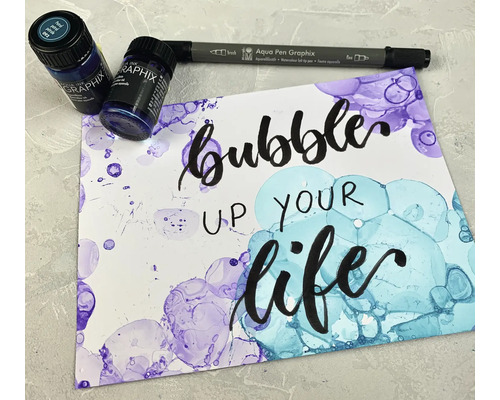 Aquarellfarben, Stift und mit Aquarellfarbe gestaltetes Papier mit dem Schriftzug Bubble up your life