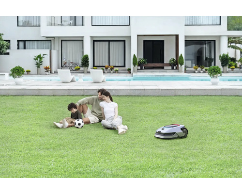 Une famille se détend sur la pelouse à côté d''un robot tondeuse devant une maison moderne avec piscine.