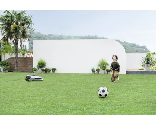 Un garçon joue au football sur une pelouse pendant qu''un robot tondeuse tond.