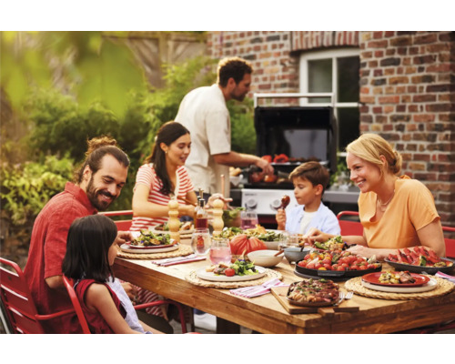 Famille appréciant un repas à table en bois dans le jardin avec barbecue en arrière-plan