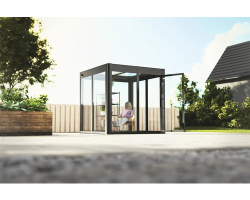 Quadratisches Glashaus mit geöffneter Tür im Garten, eine Frau sitzt auf einem Stuhl