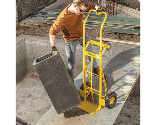 Eine Person transportiert mit einer gelben Sackkarre einen grauen Behälter auf einer Baustelle.