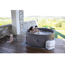 Scène avec un couple se détendant dans un spa gonflable sur une terrasse en bois. À l''arrière-plan, on aperçoit une zone résidentielle et des collines.