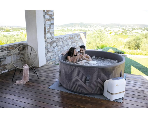 Scène avec un couple se détendant dans un spa gonflable sur une terrasse en bois. À l''arrière-plan, on aperçoit une zone résidentielle et des collines.