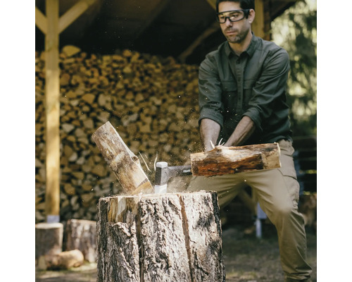 Un homme fend du bois avec une hache sur un billot, avec du bois de chauffage empilé en arrière-plan.