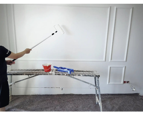 Ein Mann streicht eine Wand mit einer Farbrolle und einem Verlängerungsstiel. Auf einem Gerüst stehen ein Farbeimer und ein Farbraster.