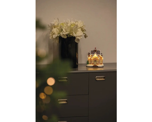 Scène décorative avec vase de fleurs artificielles et maison en pain d''épices illuminée sur une commode.