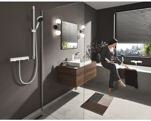 Salle de bains avec douche, lavabo et baignoire. Une femme est assise au bord de la baignoire et se détend avec une boisson.