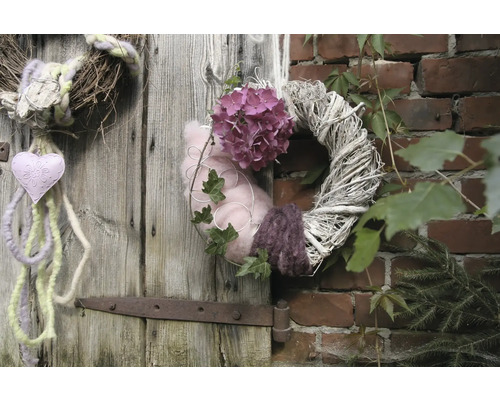 Des couronnes décoratives sont suspendues à une porte en bois et à un mur de briques, ornées de fleurs, de cœurs et de plantes.