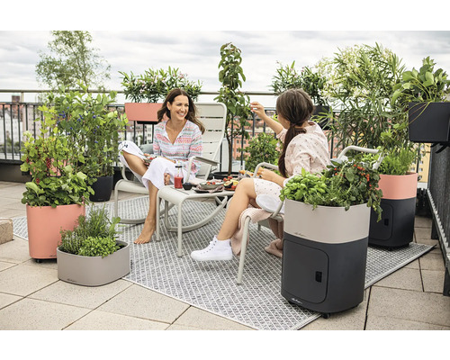 Szene auf einer Dachterrasse mit zwei Frauen, Gartenmöbeln und Pflanzgefäßen.